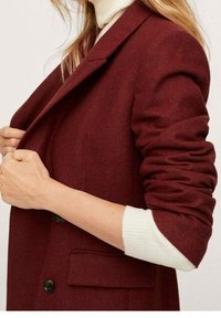 Veste en mélange de laine bordeaux avec boutons noirs, une poche poitrine unique et manches retroussées sur un pull crème clair.
