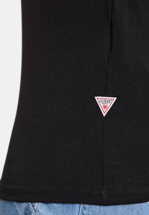 Tessuto nero con una piccola toppa triangolare bianca che presenta il testo rosso "GUESS BEACH" e un'icona di palma vicino alla cucitura inferiore.