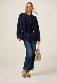Mujer llevando un abrigo navy de doble botonadura, top oscuro, jeans azules con botones dorados, tacones de piel de serpiente y sosteniendo un bolso beige.