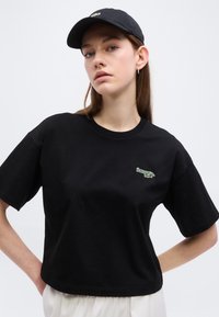 Νεαρή γυναίκα που φοράει μαύρο t-shirt Lacoste και μαύρο καπέλο, στέκεται με τα χέρια στη μέση μπροστά σε απλό φόντο.