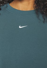 Tyrkysová mikina s kulatým výstřihem a žebrovanými detaily, s malým bílým logem Nike umístěným na středu hrudi. Měkká textura, minimalistický design.