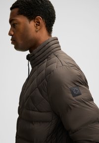 Giacca imbottita e trapuntata in marrone scuro con collo alto e cerniera, caratterizzata da una distintiva toppa con logo nero sulla spalla sinistra.