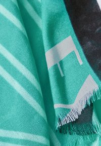 Turkos scarf med vita ränder och abstrakta mönster. Mjuk textur med fransade kanter, som visar kontrasterande svarta detaljer.