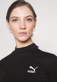 Melna ribota krekla ar garām piedurknēm, kuram ir augsta apkakle un baltas sānu svītras, ar mazu baltu Puma logo uz krūtīm. Gluda tekstūra.