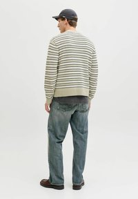 Jeune homme portant une casquette noire, un pull beige à rayures, une chemise gris foncé, un jean bleu ample et des chaussures marron, debout de dos sur un fond blanc uni.