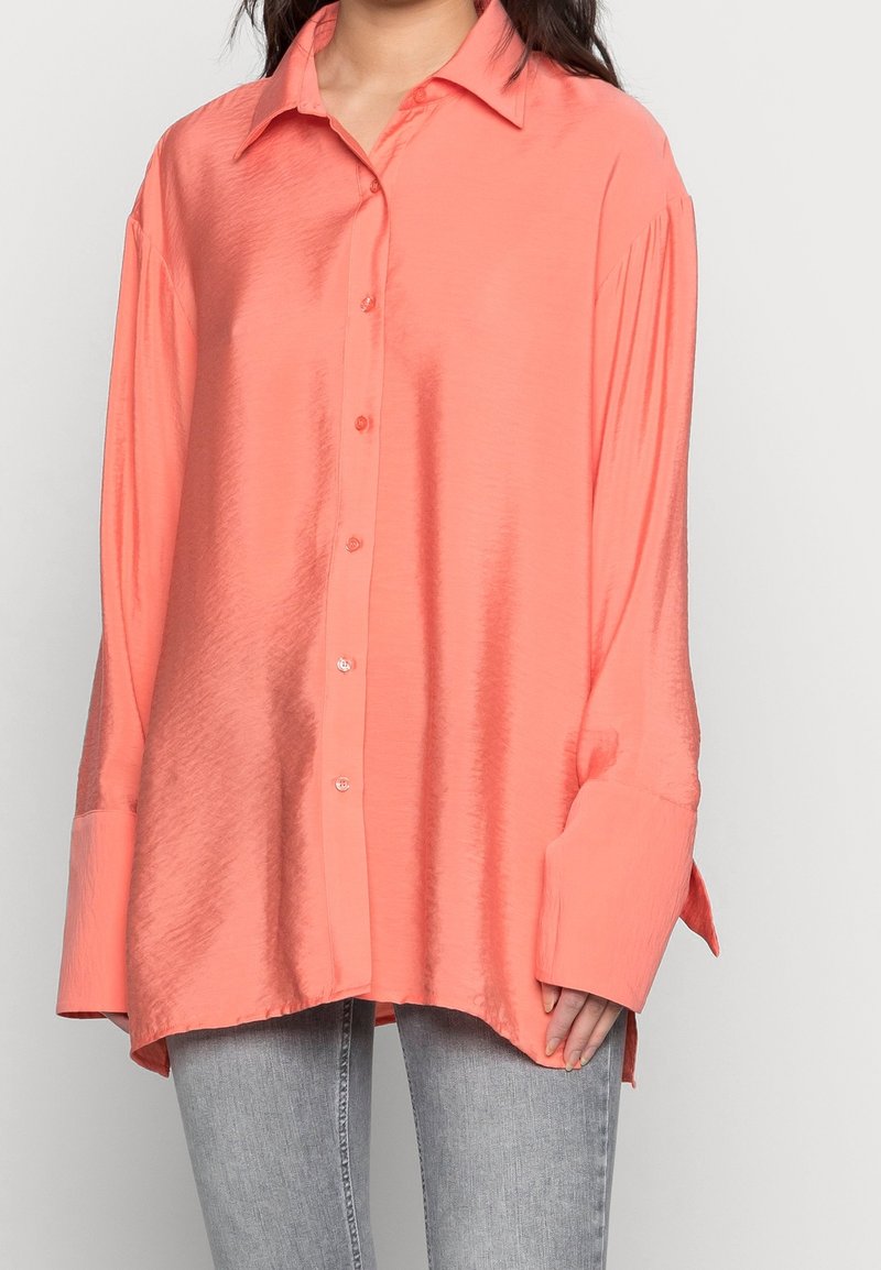 Femme portant une chemise en satin corail oversize à boutons, manches longues et larges poignets, associée à un jean gris clair.