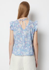 Bluse mit blauem Blumenmuster, kurzen Rüschensäumen, offener Rückseite mit Bindeschluss und geradem Saum. Leichter, strukturierter Stoff mit weißen und rosa Blumen.