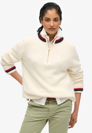 Pull en maille blanche avec une demi-fermeture éclair, doté d'un col côtelé rouge et marine. Motif décoratif aux épaules, porté sur une chemise à col.
