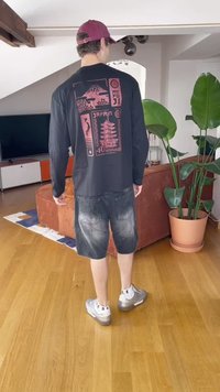 Črna majica z dolgimi rokavi, okrašena z rdečimi grafičnimi motivi Japonske, v kombinaciji z sivimi denim kratkimi hlačami in srebrnimi supergami. Notranji prostor z rastlinami.