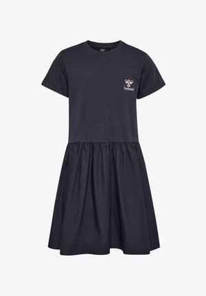 Młoda dziewczyna z kręconymi blond włosami, ubrana w czarną sukienkę z krótkim rękawem i białe trampki, stoi na jednolitym tle.