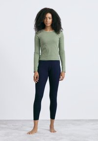 Vrouw met krullend haar staat blootsvoets op een tapijt, draagt een groene lange mouwen shirt en zwarte leggings tegen een effen witte achtergrond.