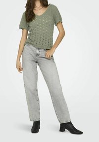 Top de punto verde oliva con escote en V y mangas cortas con borde ondulado, combinado con jeans rectos de un gris claro y botines negros de tacón.