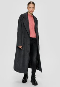 Lange grijze jas met brede revers, dubbele knoopsluiting en twee knopen. Draagt een roze blouse en zwarte jeans, gecombineerd met zwarte laarzen.
