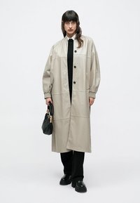 Béžový dlhý kabát s voľným strihom, s čiernymi gombíkmi, bočnými vreckami a rebrovanými manžetami, doplnený o čierne nohavice a čiernu kabelku.
