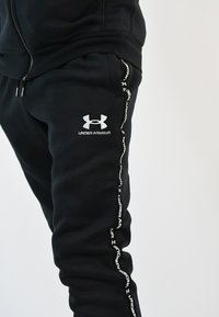 Pantalones de chándal negros con un logo blanco de Under Armour en el muslo izquierdo y un diseño de cinta blanca a lo largo de la costura lateral.