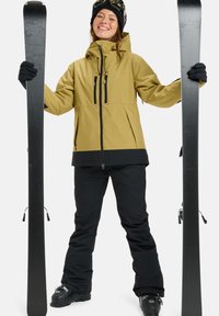 Die Skiausrüstung umfasst eine gelbe Jacke mit schwarzen Akzenten, schwarze Hosen, schwarze Handschuhe und schwarze Skier. Das Outfit hat Reißverschlüsse und eine Kapuze.