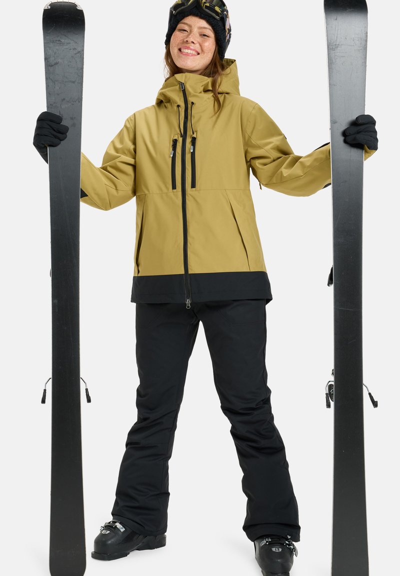 Die Skiausrüstung umfasst eine gelbe Jacke mit schwarzen Akzenten, schwarze Hosen, schwarze Handschuhe und schwarze Skier. Das Outfit hat Reißverschlüsse und eine Kapuze.
