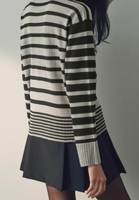 Pull rayé noir et gris avec poignets côtelés et ourlet à motifs, associé à une jupe noire. Texture douce, coupe décontractée.