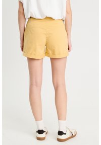 Shorts en coton jaunes avec des revers, poches plates et une texture lisse, associés à des baskets blanches et noires et des chaussettes blanches.