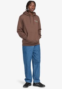 Brown hoodie with front pocket, drawstrings, and green logo detail. Paired with blue loose-fit jeans and black shoes.