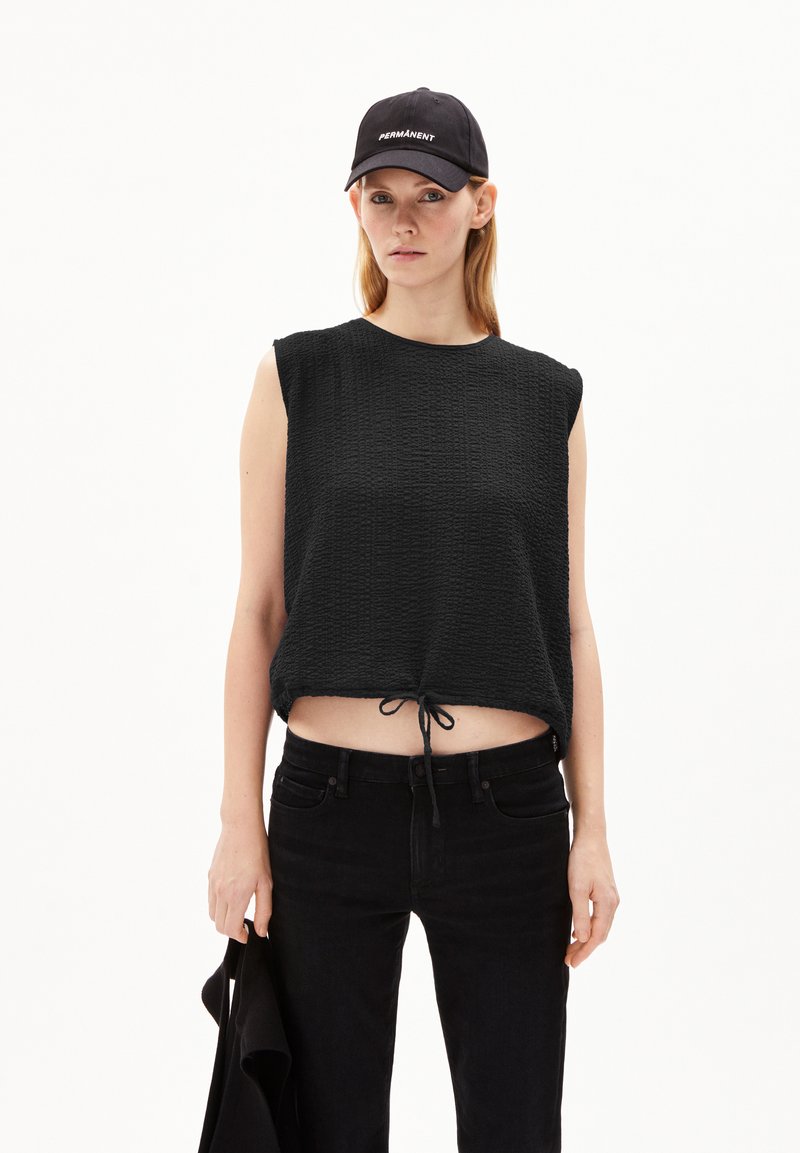 Black sleeveless top with textured fabric and drawstring hem, paired with black jeans. Black cap with white "PERMANENT" text visible.