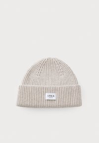 Beige strikket lue med ribbet tekstur og rullet mansjett. Har en liten rektangulær logo-lapp på forsiden. Myk stoff.