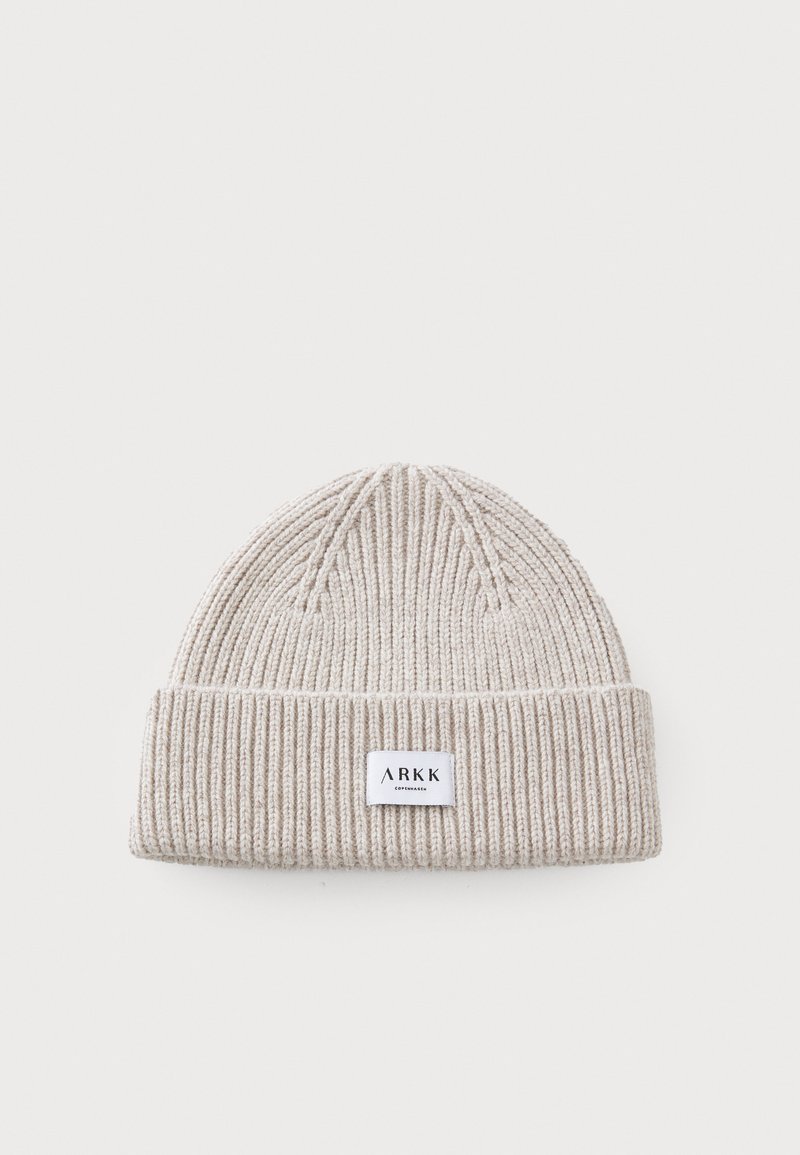 Beige strikket lue med ribbet tekstur og rullet mansjett. Har en liten rektangulær logo-lapp på forsiden. Myk stoff.