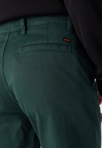 Pantalones de algodón verdes con un bolsillo trasero que presenta un sutil detalle de costura y una pequeña etiqueta de logo "BOSS" en naranja.