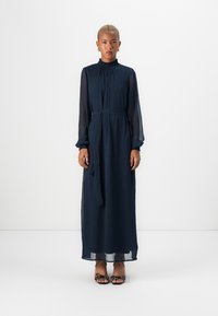 Μπλε ναυτικού, φόρεμα μακρυμάνικο με ριχτό γιακά, ζώνη στη μέση και διαφανή μανίκια. Το μοντέλο φοράει μαύρες παντόφλες με τακούνι και πουά.