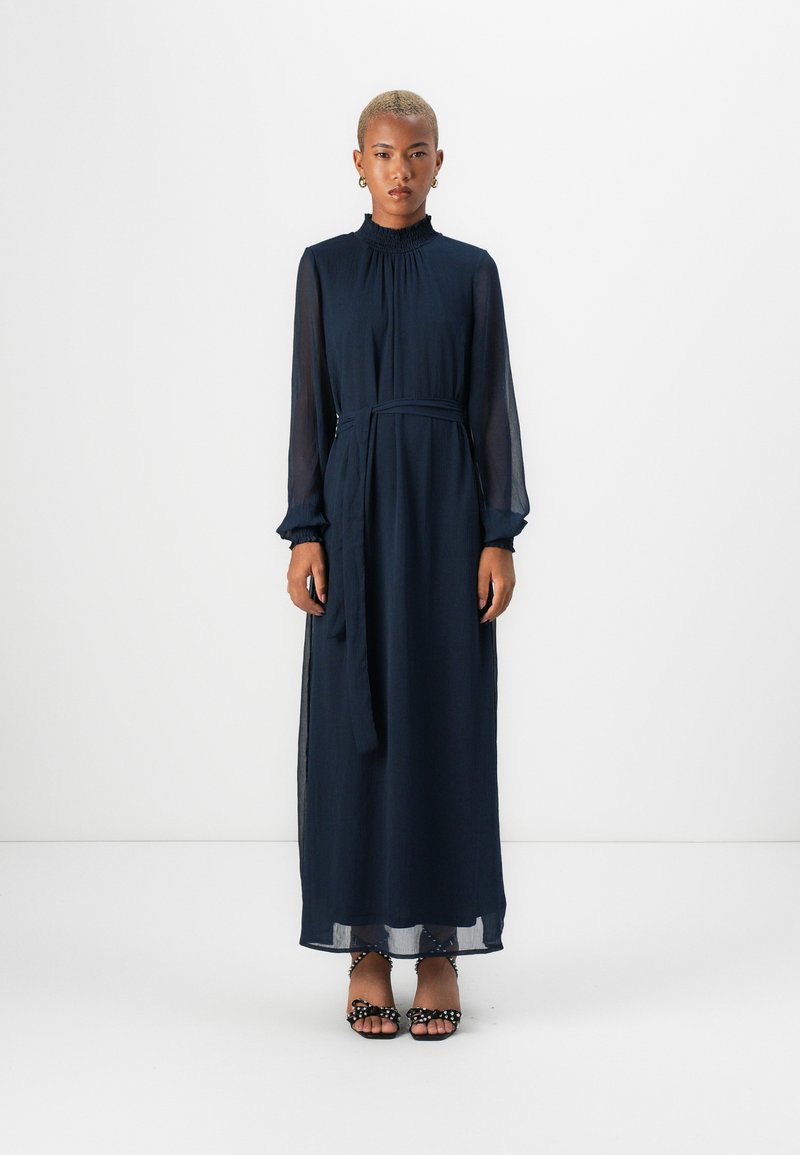 Μπλε ναυτικού, φόρεμα μακρυμάνικο με ριχτό γιακά, ζώνη στη μέση και διαφανή μανίκια. Το μοντέλο φοράει μαύρες παντόφλες με τακούνι και πουά.