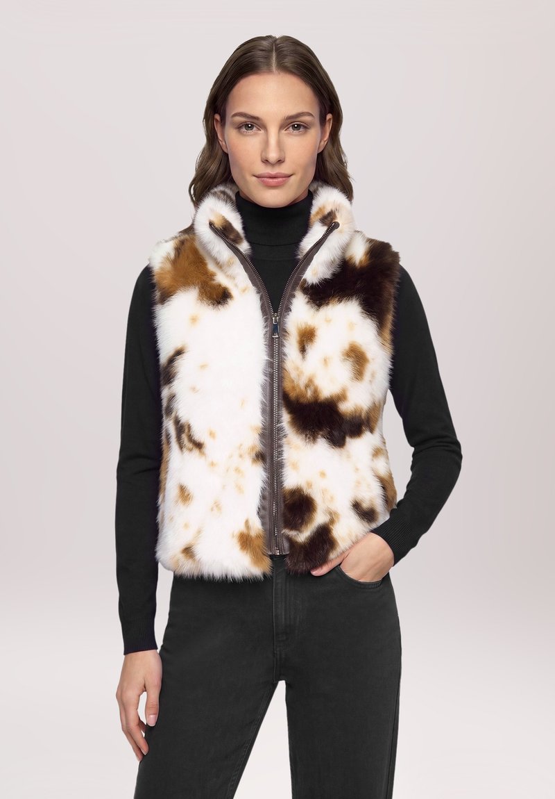 Femme aux cheveux bruns porte un gilet en fourrure sans manches blanc, marron et noir à taches, par-dessus un col roulé noir et un pantalon noir.