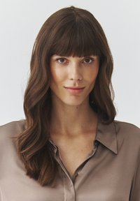 Silky brown blouse with a collar, featuring a button-down design and soft, smooth texture. Hair styled in loose waves with bangs.