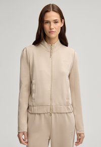 Giacca beige con zip, realizzata in tessuto morbido. Collo alla coreana, tasche frontali e discreto dettaglio del logo sul petto sinistro. Orlo aderente con zip dorata.