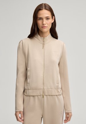 Beige Zip-Jacke mit Reißverschluss, aus glattem Stoff gefertigt. Stehkragen, Fronttaschen und dezentes Logo-Detail auf der linken Brust. Figurbetonter Saum mit goldenem Reißverschluss.