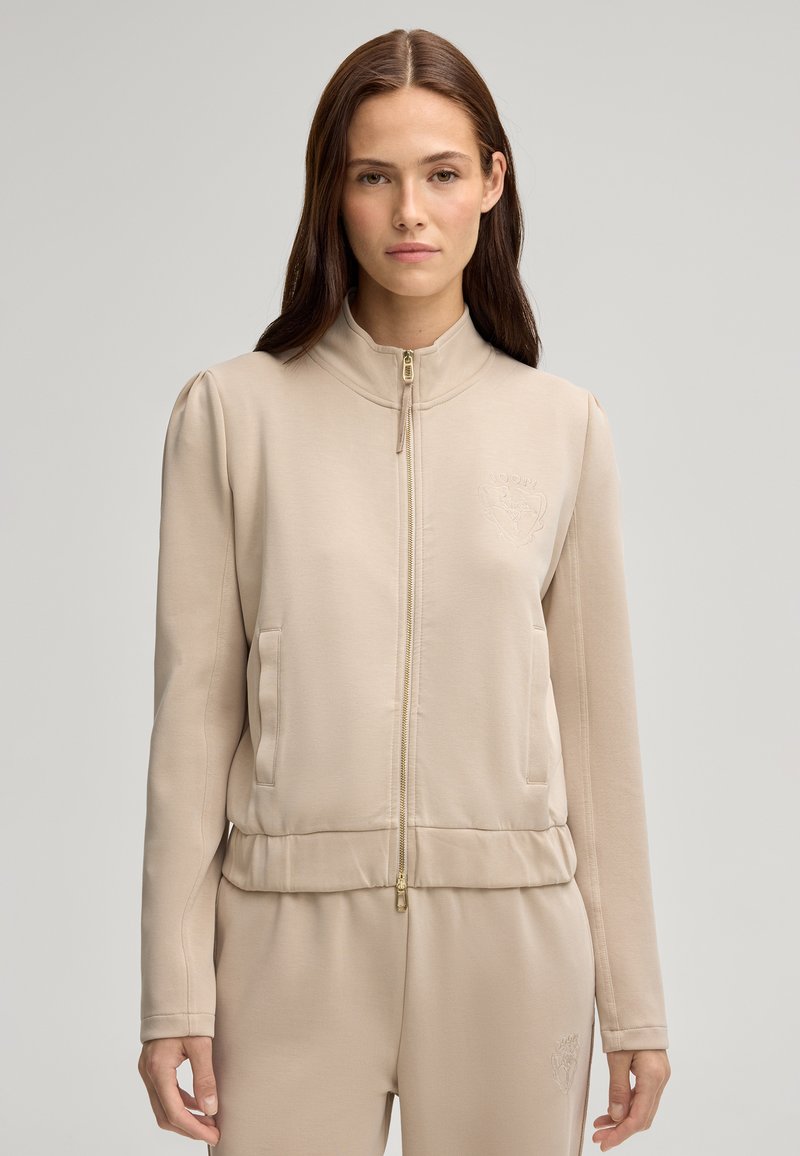 Giacca beige con zip, realizzata in tessuto morbido. Collo alla coreana, tasche frontali e discreto dettaglio del logo sul petto sinistro. Orlo aderente con zip dorata.