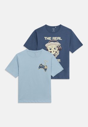 Zwei kurzärmelige T-Shirts, eines hellblau mit einem Grafikdruck von einem Spielcontroller und Keks, das andere marineblau mit einer Pizzastück-Grafik, das Kopfhörer trägt, und dem Text "THE REAL".