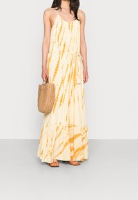 Maxi sukienka w kremowym kolorze z pomarańczowymi wzorami tie-dye, wykonana z lekkiego materiału; posiada ramiączka i sznurki w talii. W zestawie torebka pleciona.