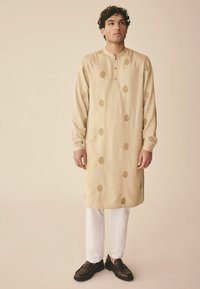 Beige Kurta mit goldenen Musterakzenten, langen Ärmeln und einem geknöpften Mandarin-Kragen, kombiniert mit weißen Hosen und dunklen Schuhen.