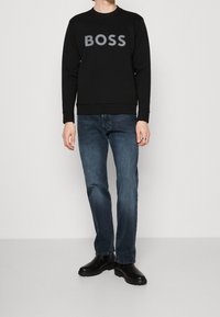 Sudadera negra con un audaz logo gris "BOSS", combinada con jeans azul oscuro y botas negras, creando un look casual y monocromático.