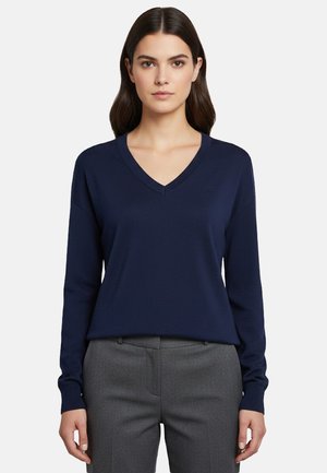 Donna che indossa un maglione a scollo a V blu navy e pantaloni grigi, in piedi davanti a uno sfondo bianco semplice.