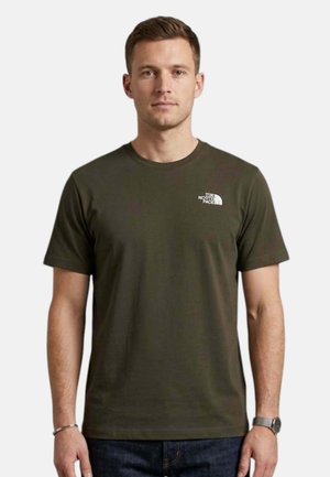 Uomo che indossa una t-shirt verde oliva The North Face e jeans scuri, in piedi davanti a uno sfondo chiaro e uniforme.
