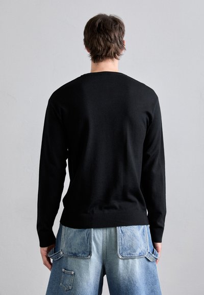 Pulover negru tricotat cu gât rotund, prezentat pe un model care poartă pantaloni scurți din denim bleu deschis cu buzunare mari. Textură netedă, mâneci lungi.