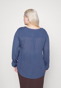 Tmavě modrá halenka s dlouhým rukávem, která má plisovaný zadní detail, kulatý výstřih a mírně zakřivený spodní lem. Lehká tkanina s jemnou texturou.