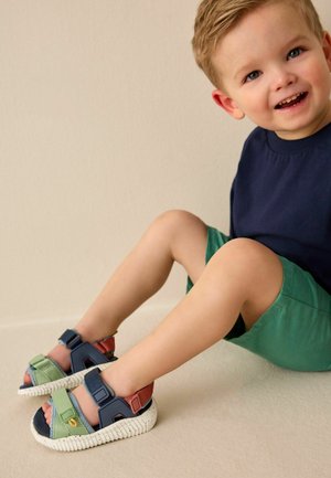 Smiling toddler wearing a navy shirt and green shorts, sitting on floor with legs extended, wearing multicolored sandals with straps.