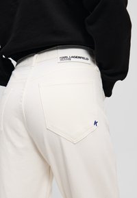 Jeans in denim bianco con una vestibilità rilassata, dotati di una tasca posteriore visibile, etichetta con il marchio "KARL LAGERFELD JEANS" e un accento ricamato blu.