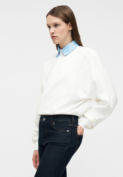 Γυναίκα που φοράει λευκό φαρδύ φούτερ με ανοιχτό μπλε γιακά, σκούρα μπλε εφαρμοστά τζιν, στέκεται με το ένα χέρι στην τσέπη, κοιτάζοντας πλάγια.