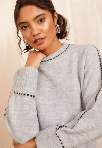 Grå sweater lavet af blødt stof med ribbede manchetter og prikket sort detaljering langs udskæringen og ærmerne. Enkel, afslappet pasform.