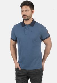 Blå polo-skjorte med marineblå krage og ermer, med et lite logo på brystet og en brun armfelg. Laget av pustende stoff.
