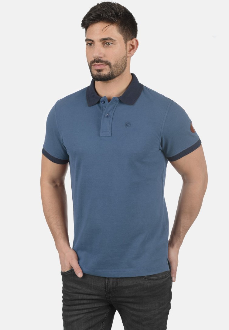Blå polo-skjorte med marineblå krage og ermer, med et lite logo på brystet og en brun armfelg. Laget av pustende stoff.