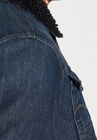 Jeansjacka i mörkblå färg med en texturerad svart fuskpäls-kant, bröstfickor och synliga sömnadsdetaljer.